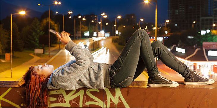 a casually dressed young female reading her phone while laying on a graffitied bridge over a roadway